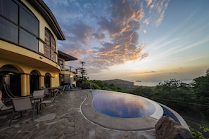 A curved infinity pool with a stone patio and outdoor seating, overlooking a scenic landscape with a view of the sea and hills. - Prime location- Two Bay spectacular view + infinity pool + 30min to LIR airport (Playa Hermosa)
