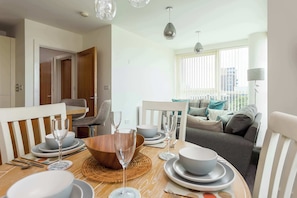 A dining area with a wooden table set for four, a sofa in the background, and a view of the city through large windows.
