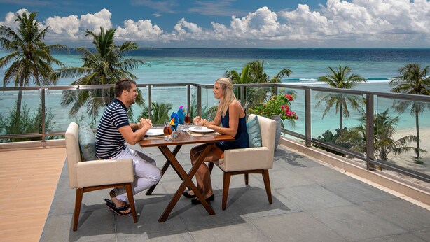 A dining table with two people seated, overlooking a beach with palm trees and the ocean. - Seasalter Maldives (Hulhumalé)