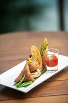 Un plato blanco rectangular con tres pastas triangulares y un vaso de bebida roja sobre una mesa de madera.