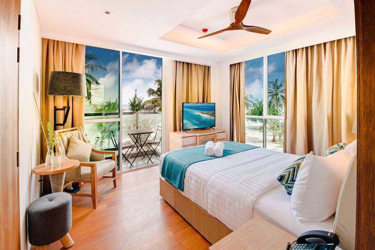 A hotel room with a bed, a TV, a chair, a table, and a balcony with a view of the ocean.