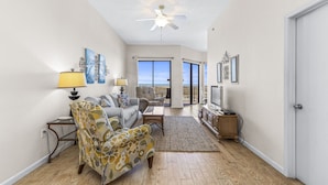 A spacious living room with a sofa, armchair, and coffee table, a ceiling fan, and a view through large windows. - Phoenix VII-111 | Beachfront | Resort Style Amenities | My Beach Getaways (Orange Beach)