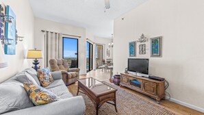 A living room with a sofa, armchair, and a coffee table. There is a television on a wooden cabinet, and a large window with curtains. - Phoenix VII-111 | Beachfront | Resort Style Amenities | My Beach Getaways (Orange Beach)