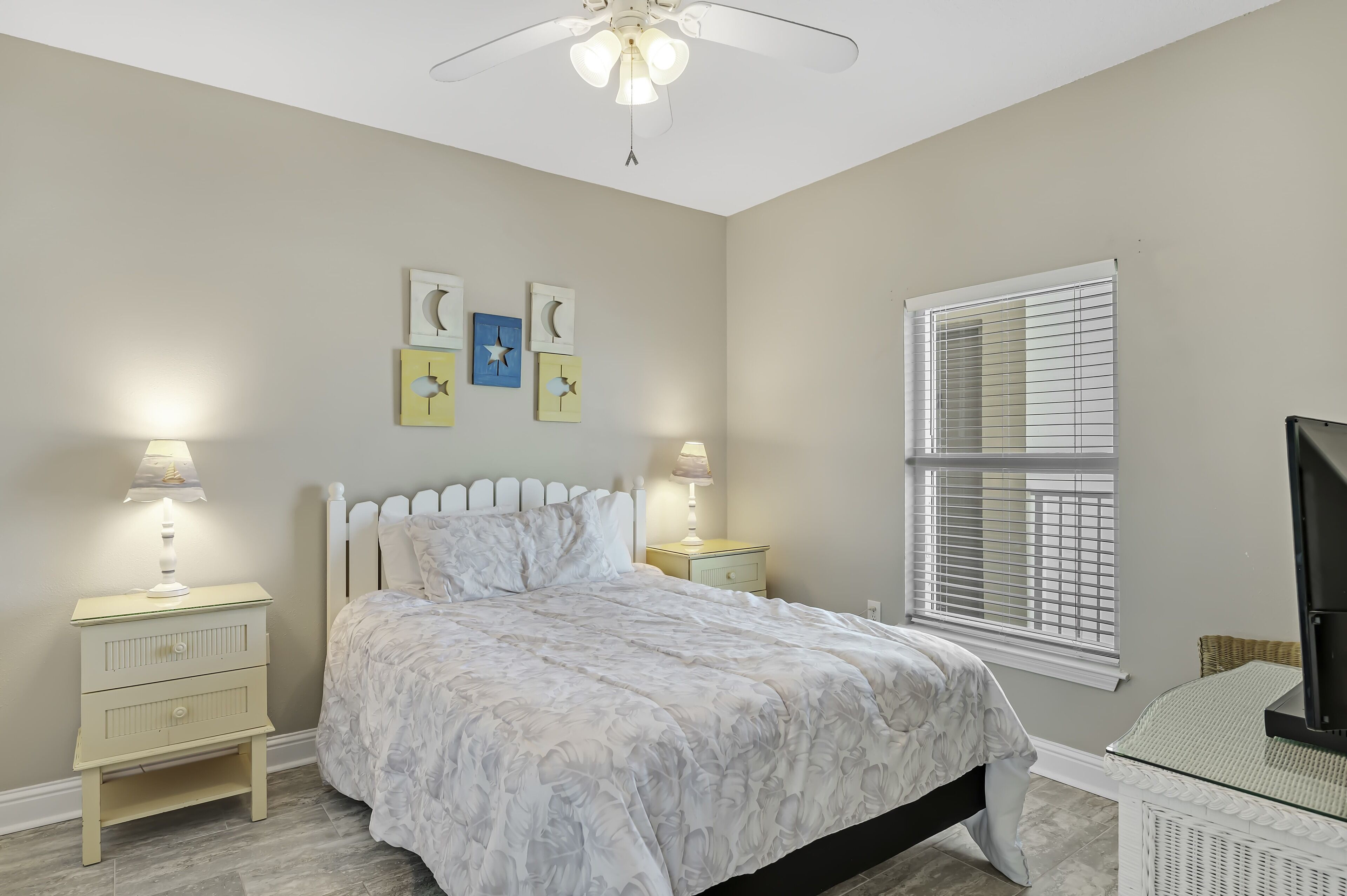 Une chambre à coucher comprenant un lit, deux tables de chevet avec des lampes, un ventilateur de plafond, une fenêtre avec des stores et des œuvres d’art murales.