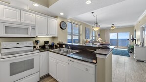 Une cuisine avec des meubles blancs, un four à micro-ondes, une cuisinière et un lave-vaisselle. Il y a une horloge au mur et, grâce à de grandes fenêtres, on peut admirer la vue sur l’océan.