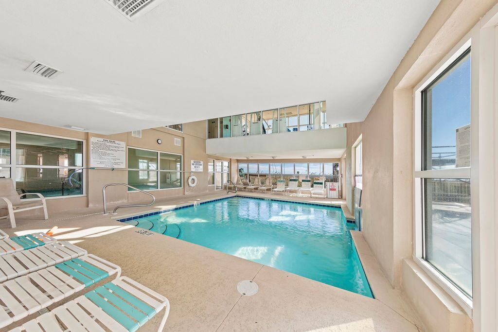 An indoor swimming pool with lounge chairs, a lifeguard stand, and a view of the city through large windows.