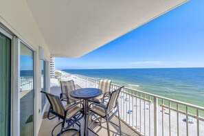Un balcon avec une table et des chaises donnant sur une plage parsemée de parasols, et offrant une vue dégagée sur l’océan.