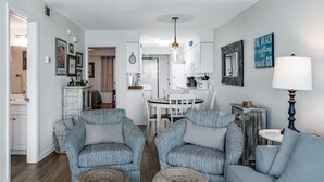 A modern living room with two grey armchairs, a sofa, and a coffee table. There is a dining area with a white table and chairs, and a kitchen in the background.