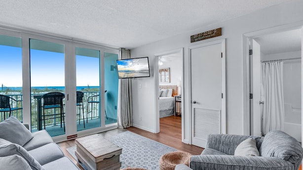 A modern living room with a large sliding glass door leading to a balcony with a view of the ocean. There is a flat-screen TV mounted on the wall, a gray sofa, and a wooden coffee table.