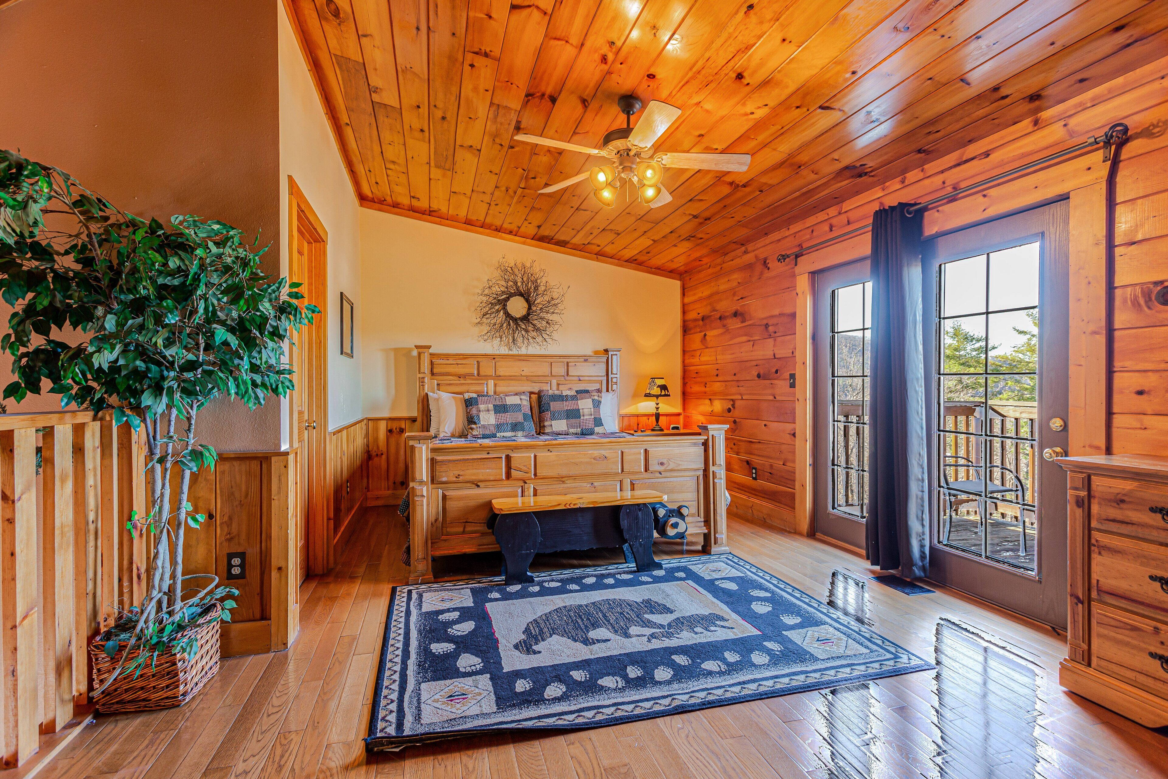 A wooden cabin interior with a bed, a ceiling fan, a large window with curtains, and a wooden dresser.