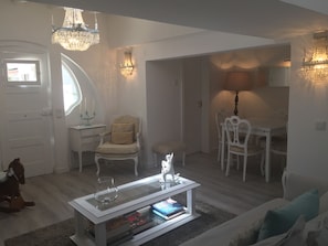 Uma sala de estar moderna com um sofá branco, uma mesa de centro de vidro com livros, uma área de jantar com uma mesa e cadeiras brancas, e um candelabro.