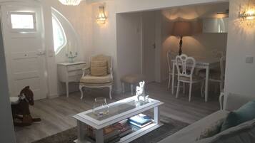 Uma sala de estar moderna com um sofá branco, uma mesa de centro de vidro com livros, uma poltrona bege e uma área de jantar com cadeiras e mesa brancas.