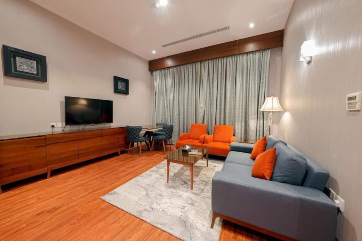 Ein modernes Wohnzimmer mit Flachbildfernseher, Holzschrank, orange-blauem Sofa und einem Essbereich mit Stühlen.