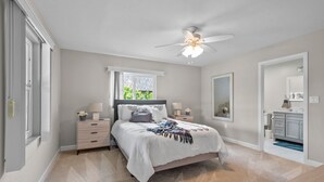 Camera da letto con un letto, comodini, una ventola a soffitto, una finestra e un bagno con uno specchio e pensili.