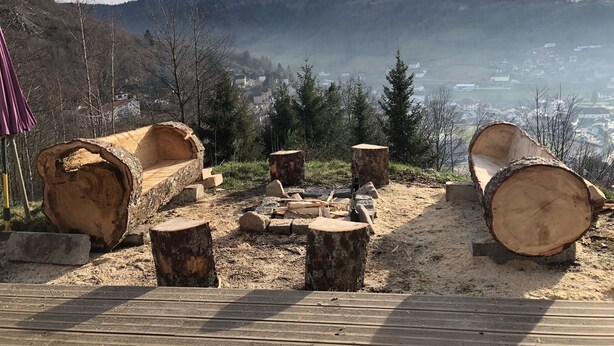 Une terrasse en bois avec des bancs en rondins et un foyer, offrant une vue sur une vallée parsemée de maisons et d’arbres.