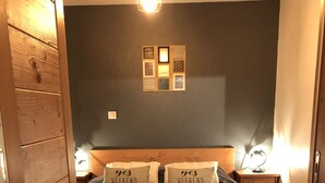 Une chambre avec un lit en bois, une couvre-lit bleu foncé, deux oreillers ornés de l’inscription « oui, week-end », un luminaire fixé au mur et une petite table de chevet en bois avec une lampe.
