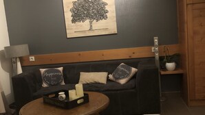 Un salon avec un mur de couleur gris foncé, une tête de lit en bois, un canapé noir agrémenté de coussins à motifs, une petite table basse ronde en bois, un petit tabouret blanc et un tableau représentant un arbre.