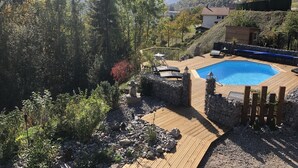 Une terrasse en bois mène à une piscine rectangulaire bordée d’un muret en pierre et d’une clôture en bois.