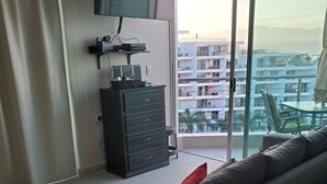 A modern living room with a grey sofa, a flat-screen TV mounted on the wall, a small cabinet, and a balcony with a view of a residential area.