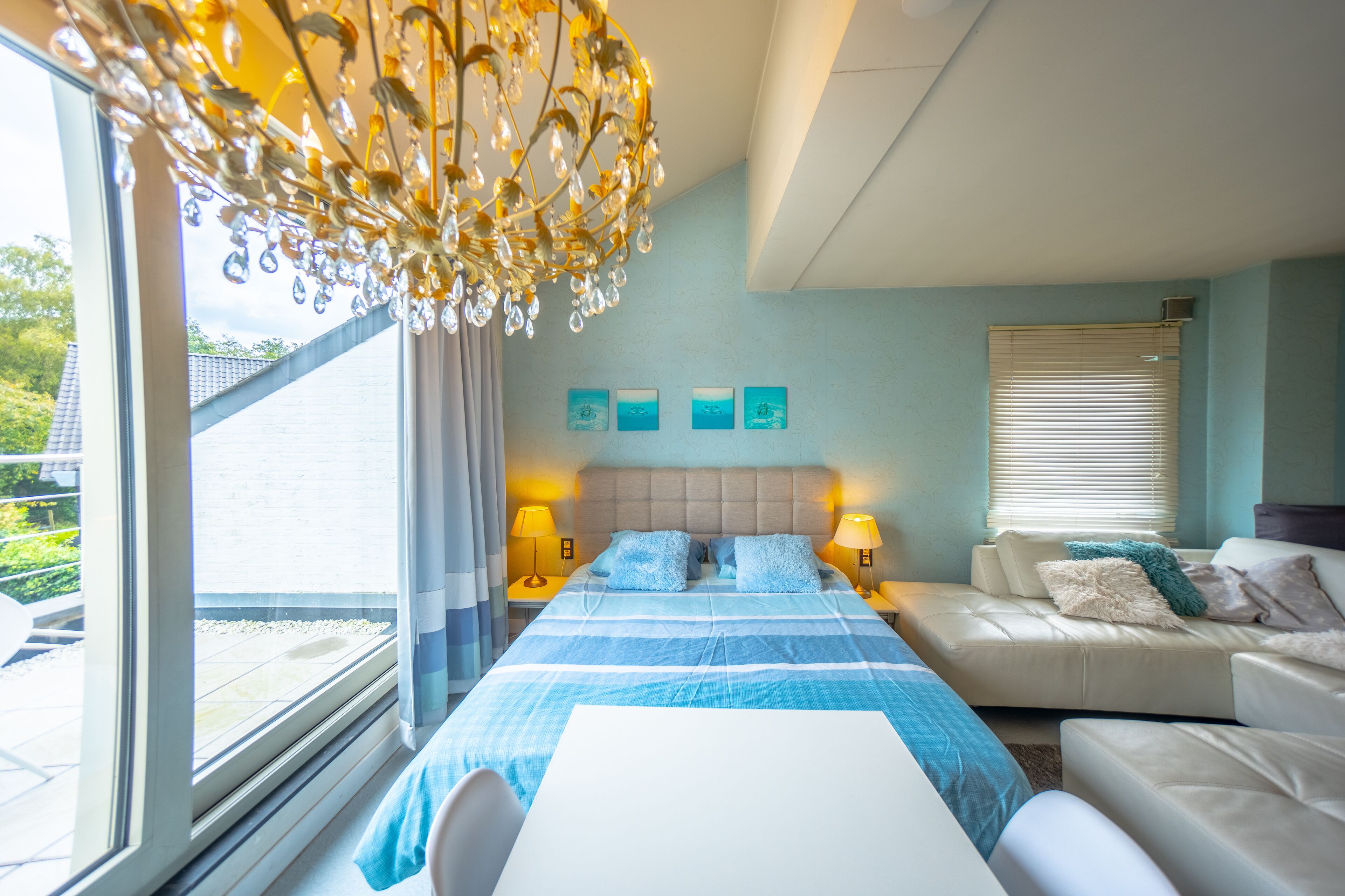 A hotel room with a large bed, a sofa, and a dining table. There are three paintings on the wall and a chandelier above the bed.