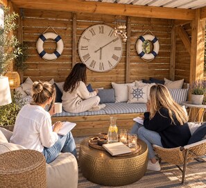 Eine hölzerne Sitzgruppe im Freien mit einer großen Wanduhr, Kissen und einem runden Tisch mit Kerzen.