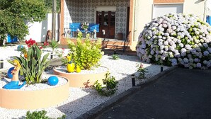 Une maison avec un toit en tuiles, une véranda avec des chaises bleues et un grand parterre fleuri de fleurs blanches.