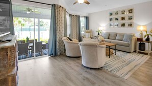 Una sala de estar moderna con un sofá, sillones, una mesa de centro y una televisión. Hay un balcón con vistas a zonas verdes.