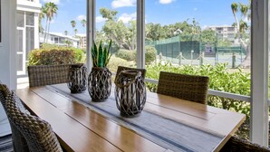 Comedor con mesa de madera y sillas de mimbre, con vista a una cancha de tenis y follaje.