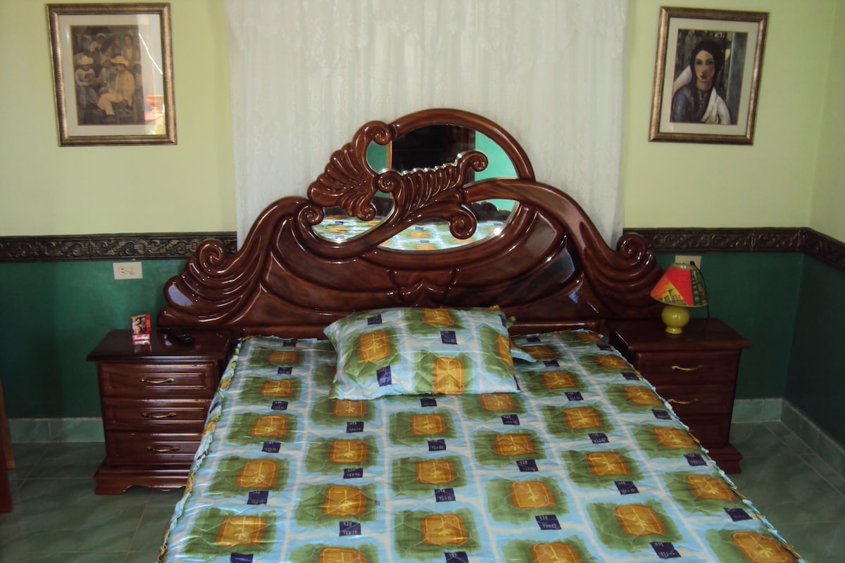A bedroom with a wooden bed frame, a patterned bedspread, two bedside tables with lamps, and framed pictures on the walls.