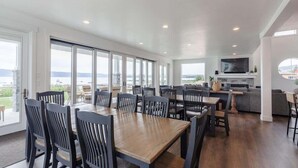 Un comedor amplio con una gran mesa y sillas, una zona de cocina con televisor, y ventanas grandes que ofrecen vistas al exterior.