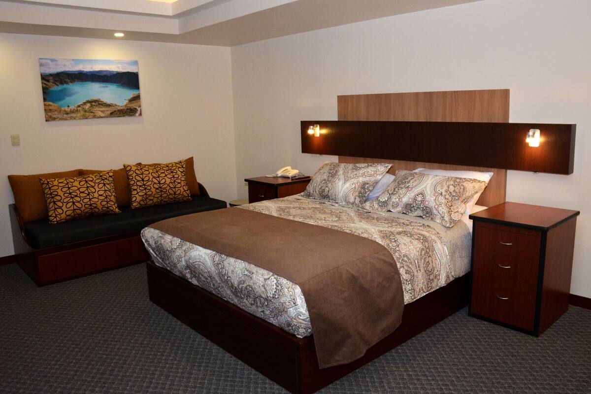 A hotel room with a large bed, a sofa, a bedside table, and a wall-mounted lamp.