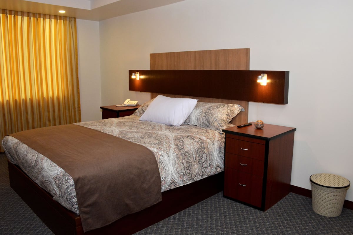 A hotel room with a large bed, wooden headboard, bedside tables, a waste basket, and a curtain.