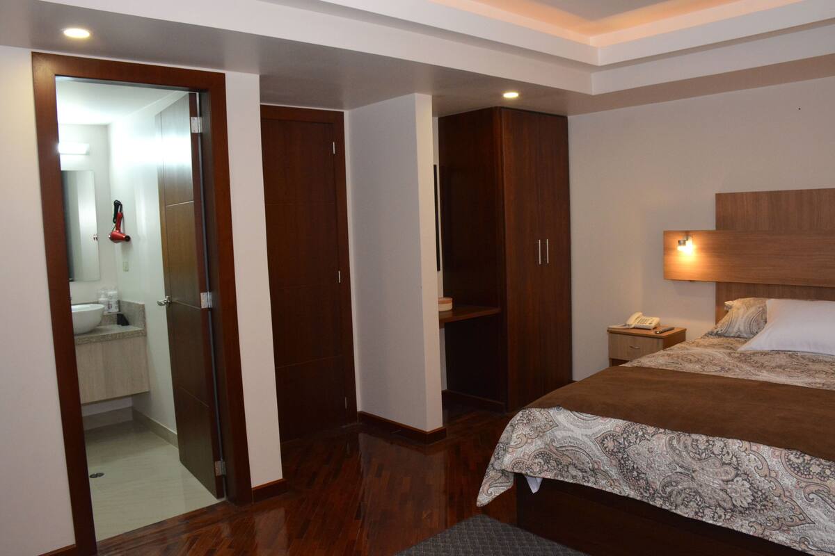A bedroom with a bed, a nightstand, a bedside lamp, a wardrobe, and a bathroom visible through an open door.