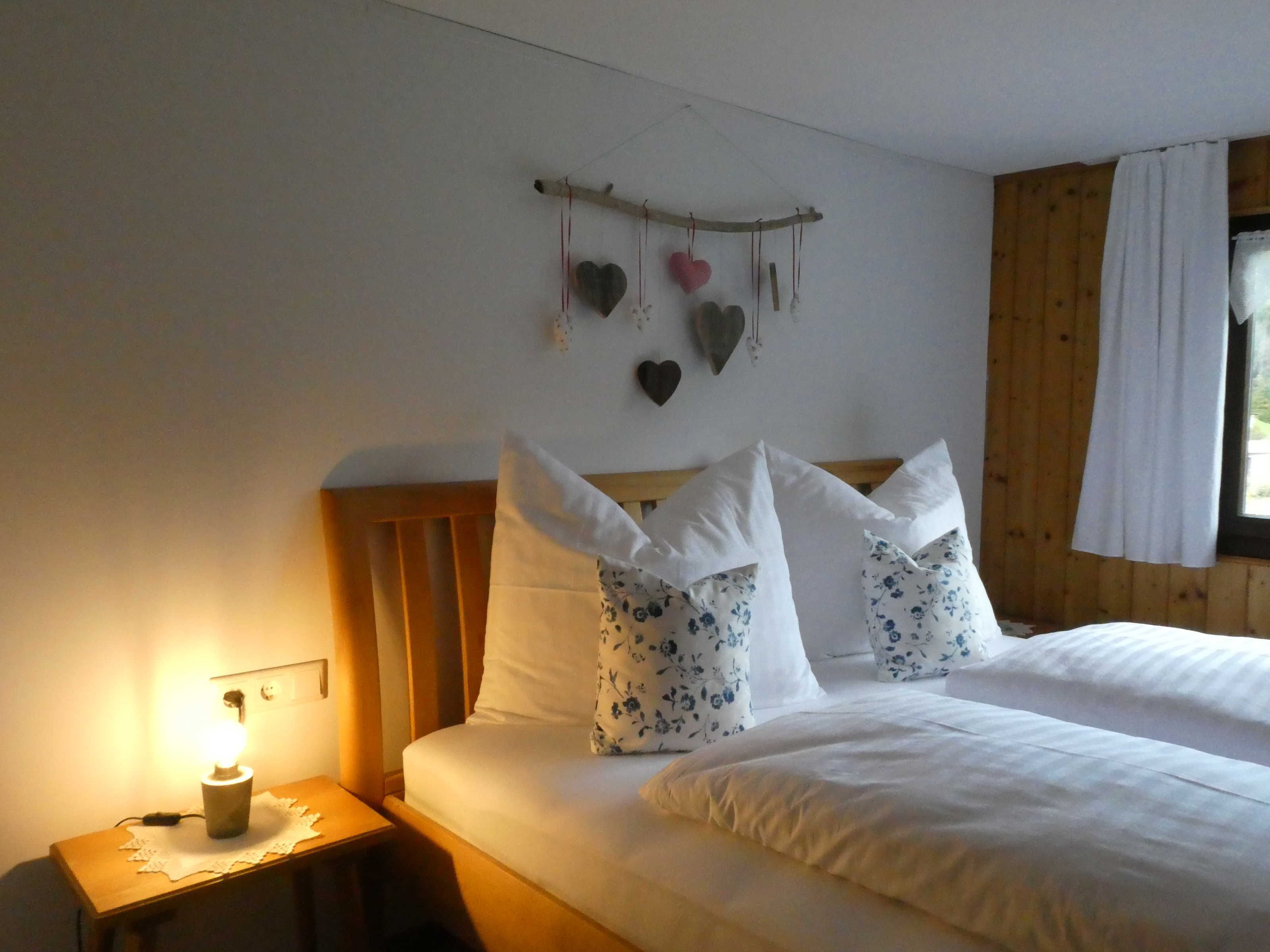 A bedroom with a wooden headboard, white bedding, floral pillows, a bedside lamp, and heart-shaped decorations on the wall.