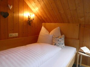 A wooden room with a bed, pillows, a wall-mounted light, and a small table with a book.