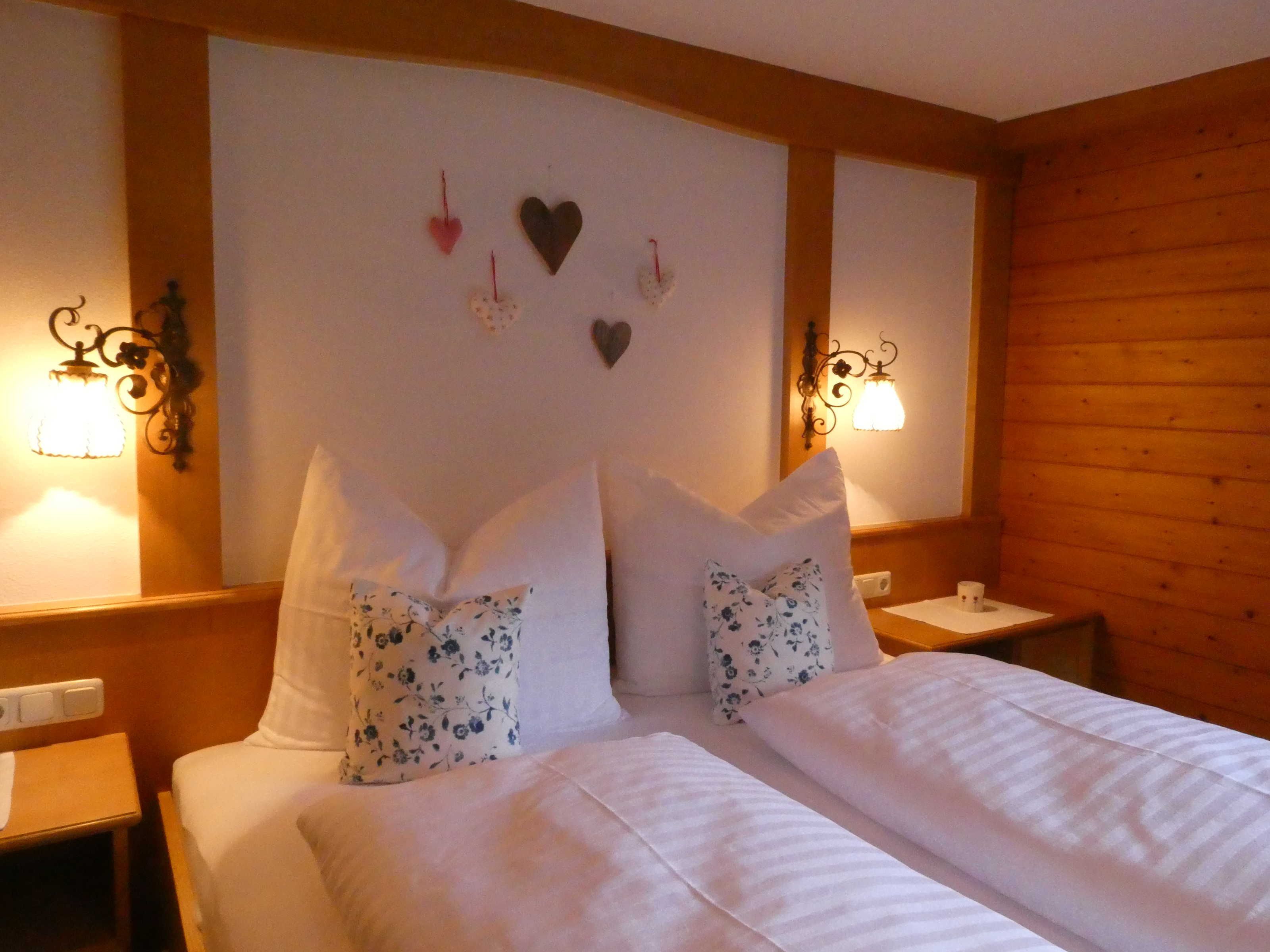 Un dormitorio con cabecera de madera, dos mesitas de noche, una cama con ropa de cama blanca y almohadas florales, y luminarias fijadas a la pared.