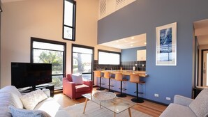 A modern living room with a TV, a red armchair, a glass coffee table, and a kitchen area with bar stools. - Cove View Retreat St.Helens (St Helens)
