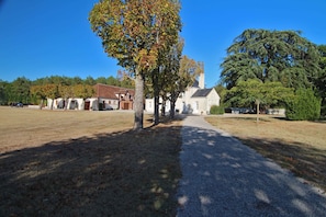 A rural setting with a gravel path leading to a white building with a bell tower, surrounded by trees and open fields.