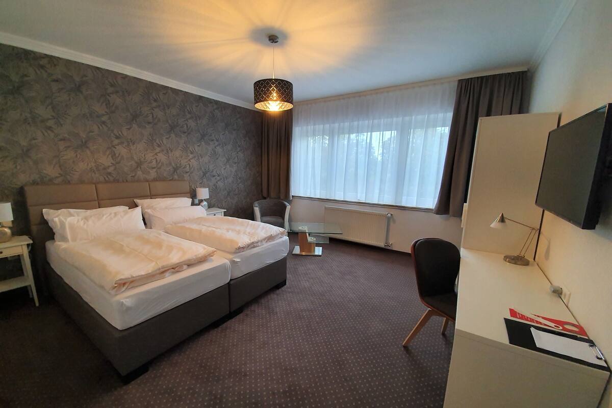 A hotel room with a double bed, a desk with a chair, a TV, and a window with curtains.