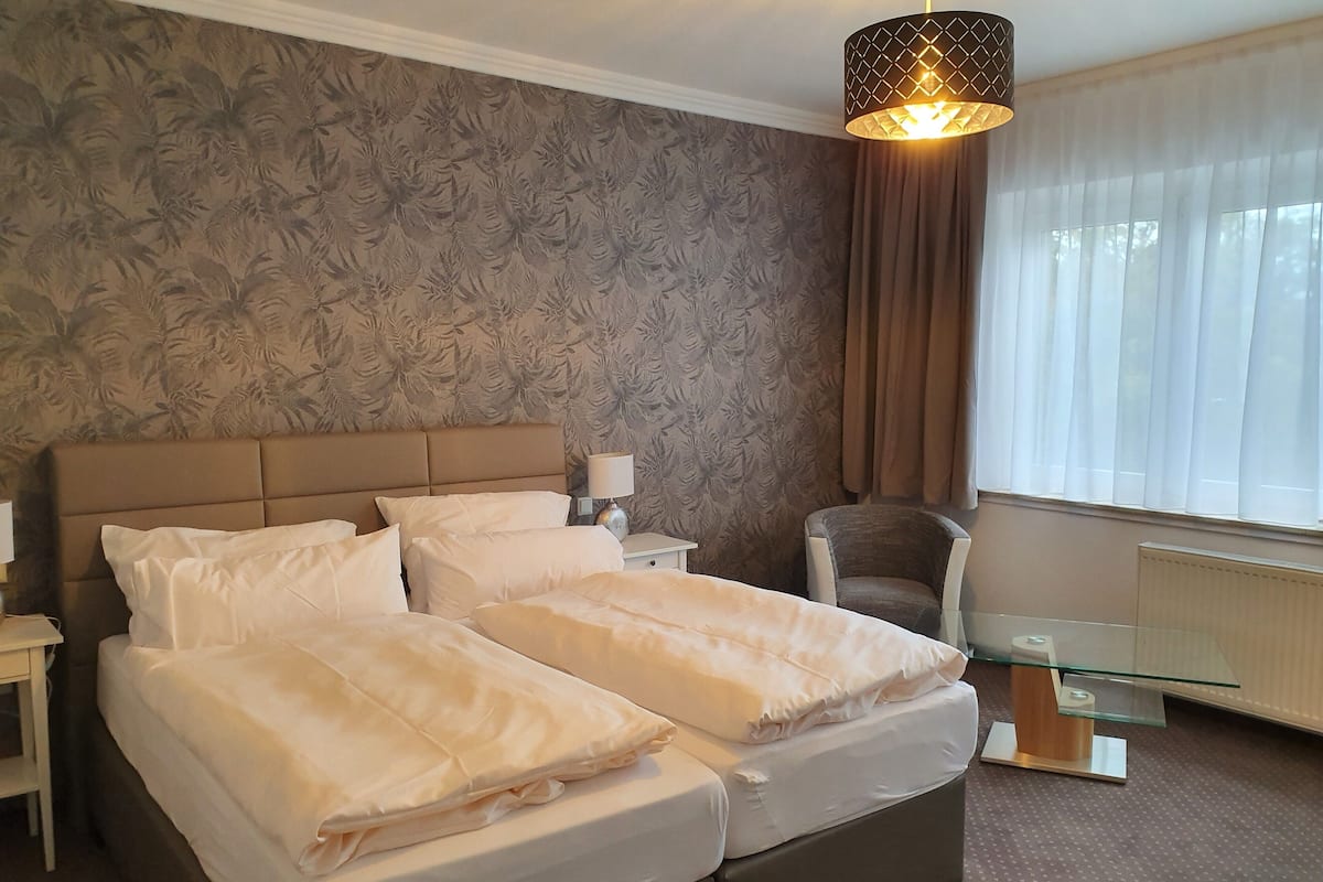 A hotel room with a large bed, a chair, a glass table, and a window with curtains.