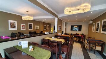 A hotel dining area with multiple tables and chairs, a reception desk, and a television screen.