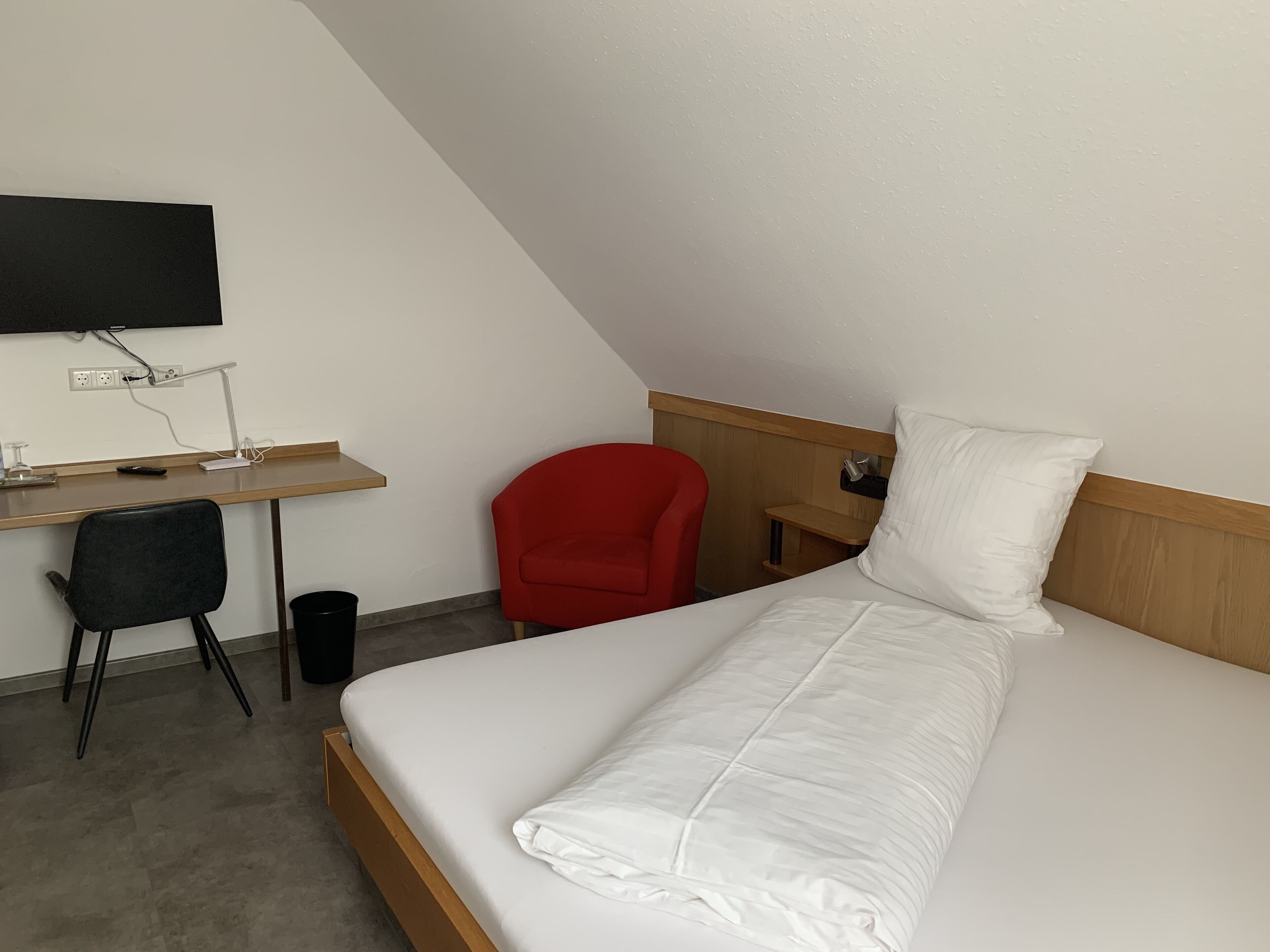 Una habitación pequeña y moderna con una cama, un escritorio, una silla, un televisor y un cesto de basura.
