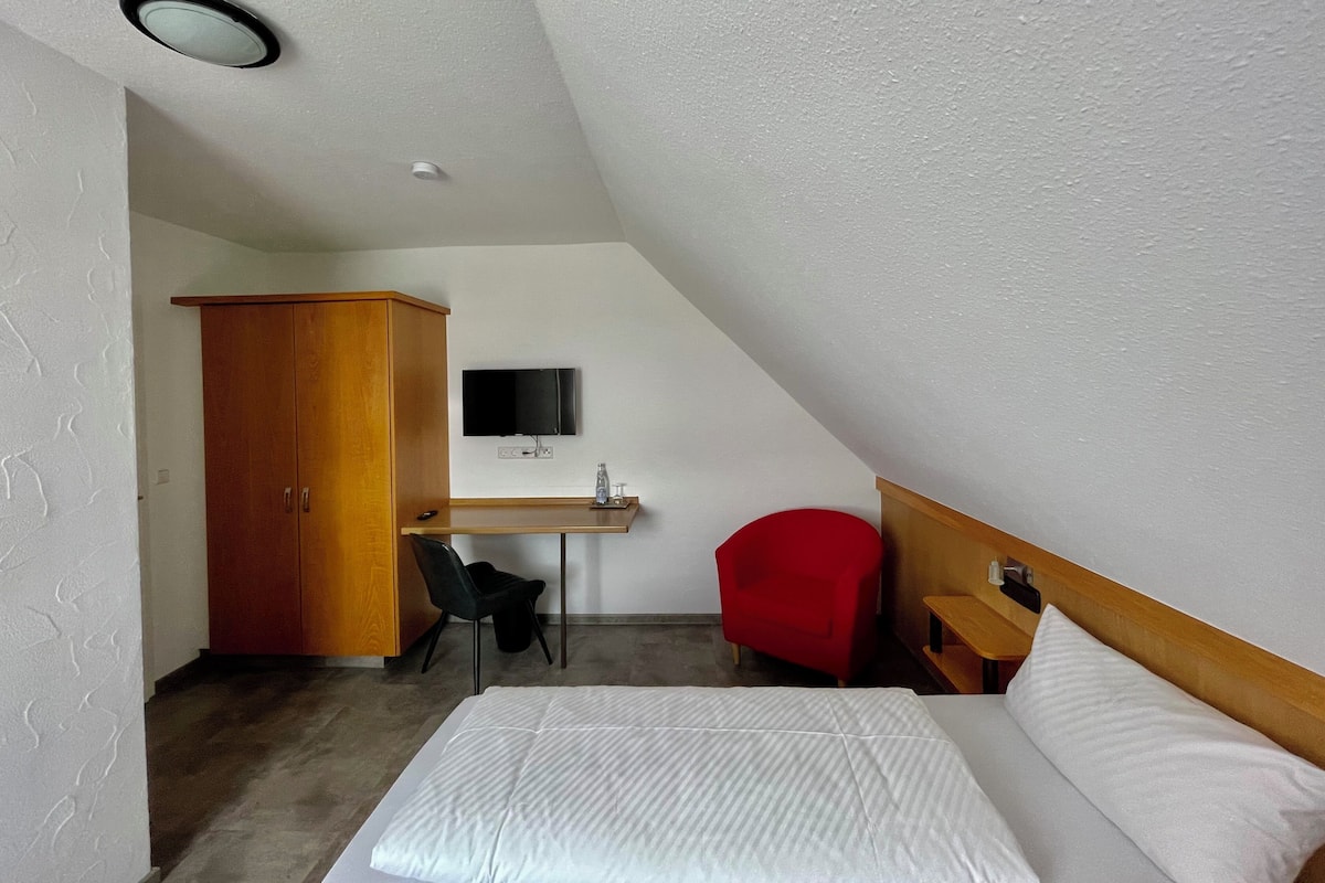 Um quarto com uma cama, uma escrivaninha com televisão, uma cadeira vermelha e um guarda-roupa.