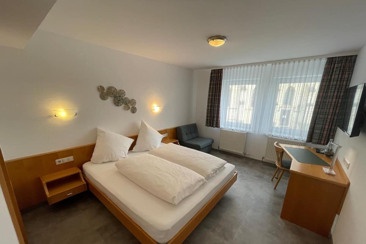 Quarto de hotel com cama, escrivaninha, cadeira, televisão e janela com cortinas.