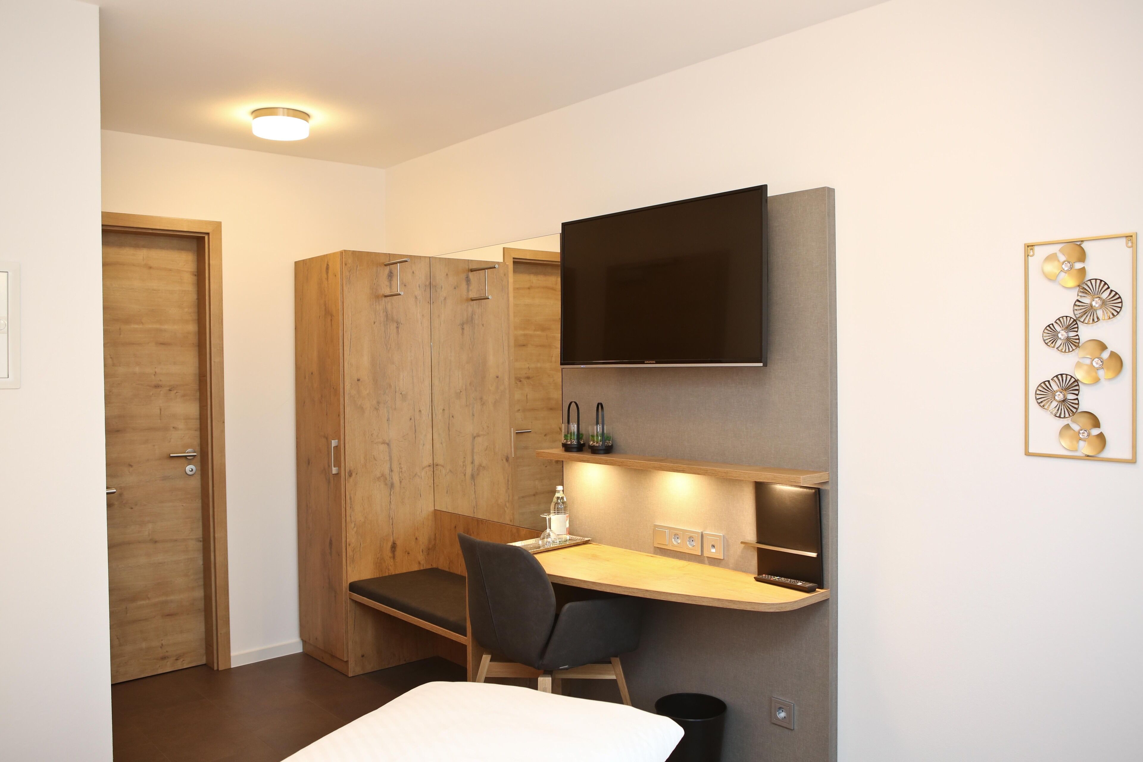 A compact hotel room with a wooden wardrobe, a desk with a chair, a flat-screen TV mounted above the desk, and a wall-mounted artwork.