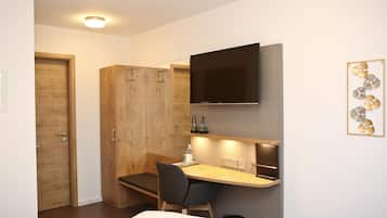 A compact hotel room with a wooden wardrobe, a desk with a chair, a flat-screen TV mounted above the desk, and a wall-mounted artwork.