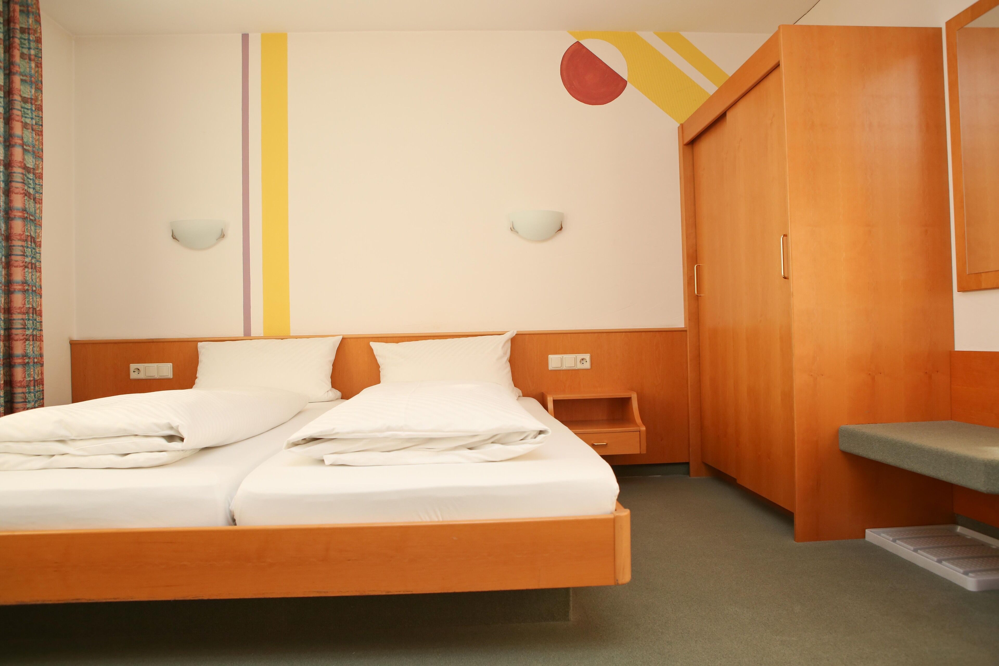 A hotel room with a wooden bed, a wardrobe, a bedside table, and a wall with a geometric design.