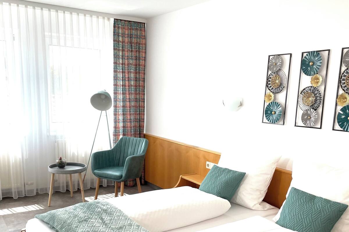 Quarto de hotel com uma cama grande, duas cadeiras, uma mesa pequena e uma janela com cortinas.