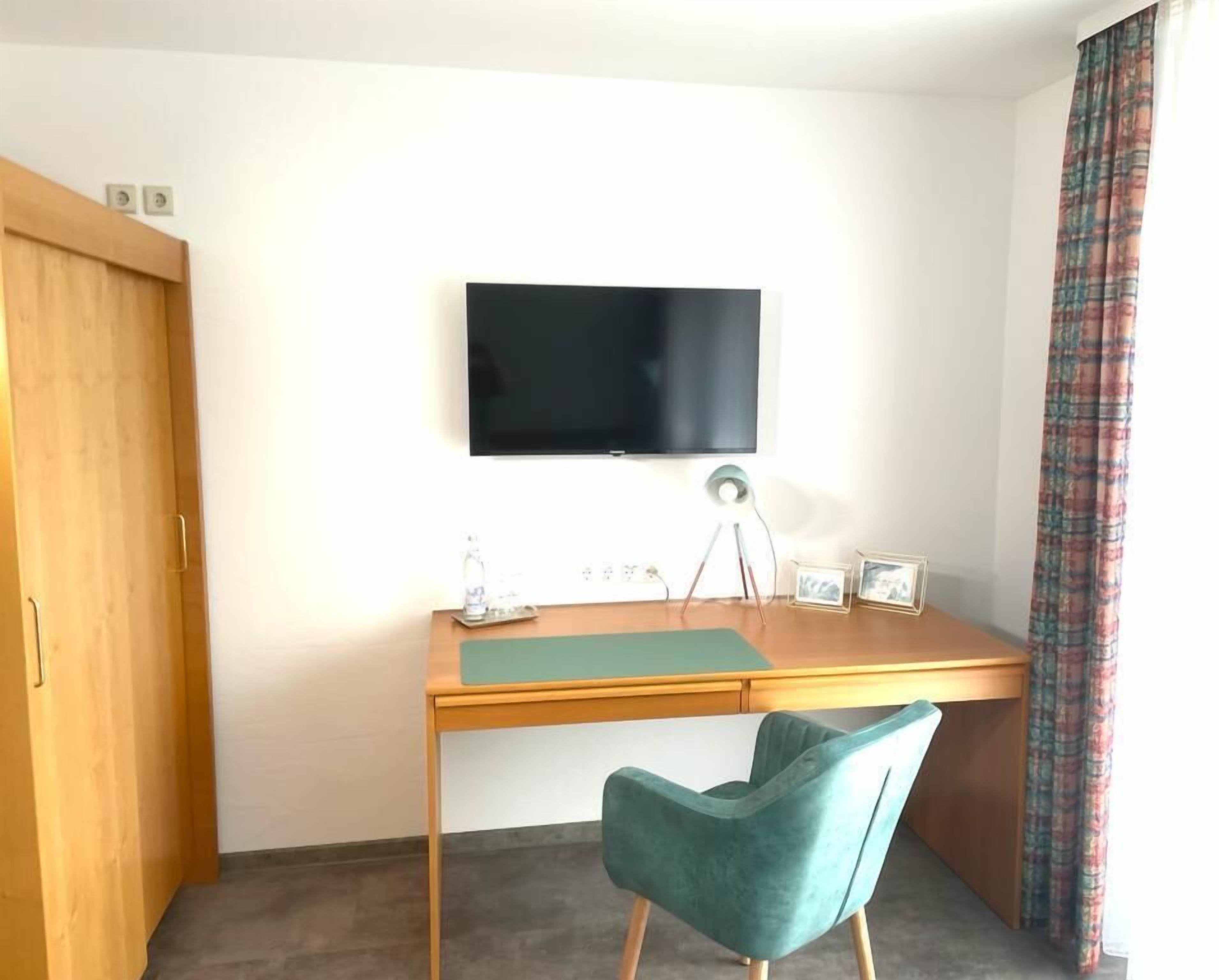 Quarto com escrivaninha de madeira, cadeira verde-água, televisão de tela plana na parede e guarda-roupa de madeira.
