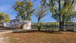 Una casa de un piso con porche y una pequeña terraza, cerca de un lago rodeado de árboles y una mesa de picnic.